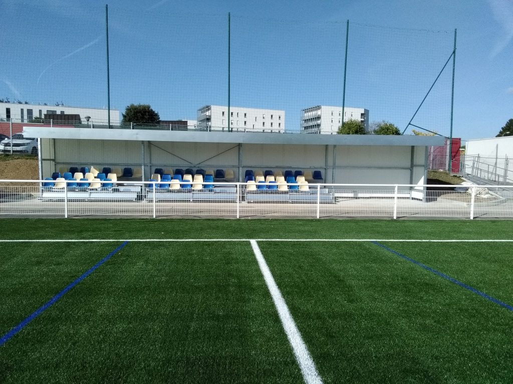 Spectator shelter with blue and white seats seen from the artificial pitch