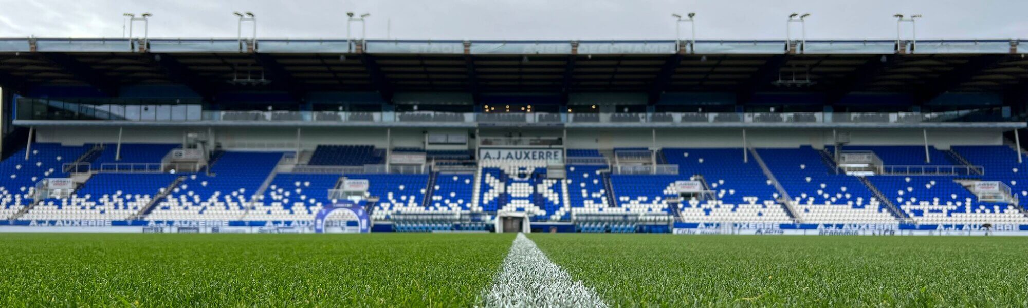 Façade de tribune sous ciel couvert, sièges bleus et blancs dessinant le blason de l'AJ Auxerre