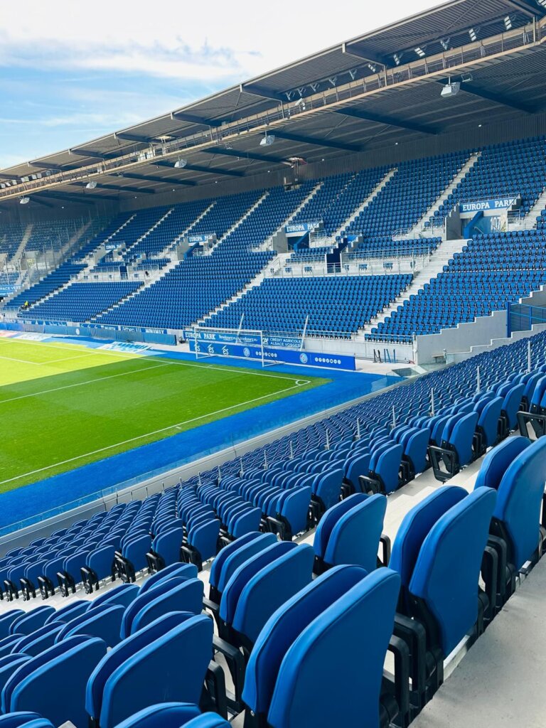 Low-angle view of a stand at the Meinau, tiers filled with blue seats from top to bottom beneath the roof structure