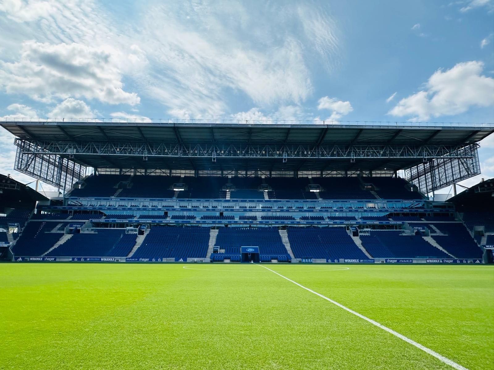 Tribune principale du Stade de la Meinau vue depuis la pelouse, deux niveaux de gradins équipés de sièges bleus